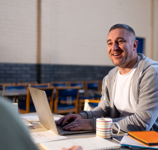 A man smiling and talking to someone while using a laptop in a community centre
